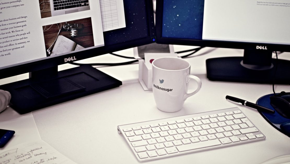бяла керамична чаша между магическата клавиатура на Apple и два компютърни монитора с плосък екран