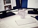 бяла керамична чаша между магическата клавиатура на Apple и два компютърни монитора с плосък екран